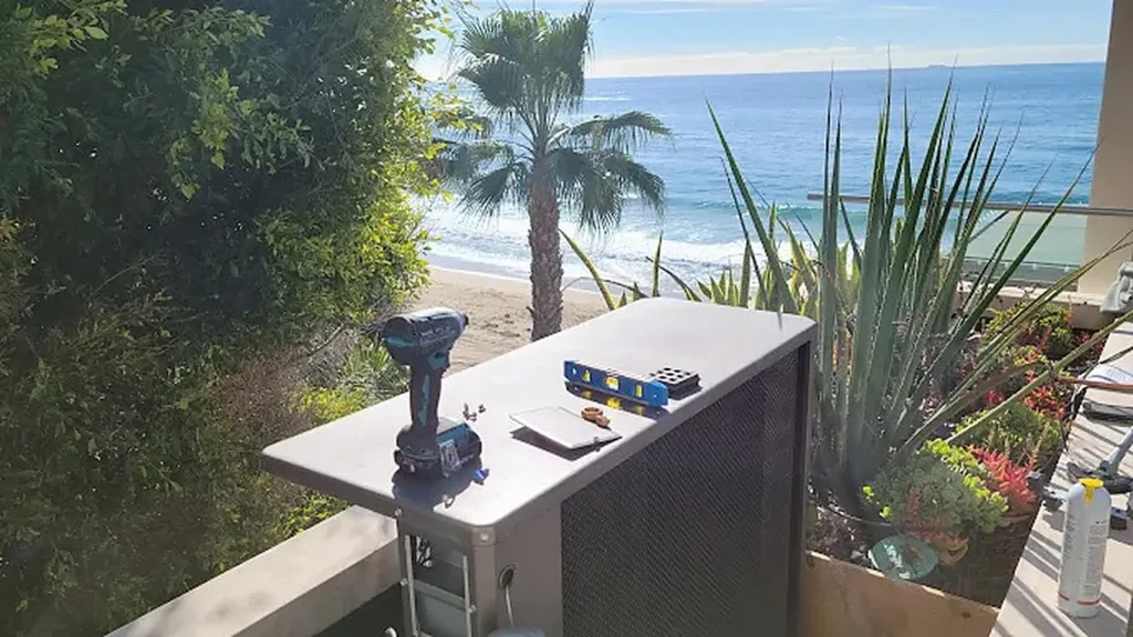 Brightwater heat pump outdoor unit being installed on an oceanfront deck in coastal Orange County, with the beach and Pacific Ocean directly behind it